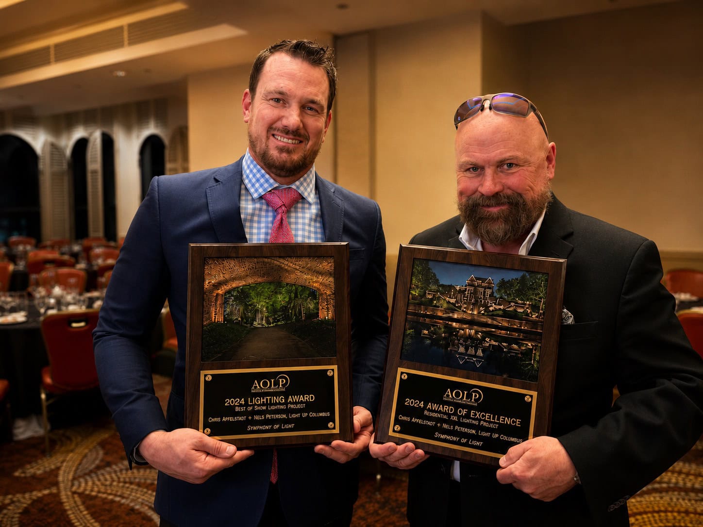 Two men in suits stand indoors, each holding a wood-framed award plaque with photos and the year 2024, smiling at the camera in a banquet hall setting. - Christopher A. Lighting Design