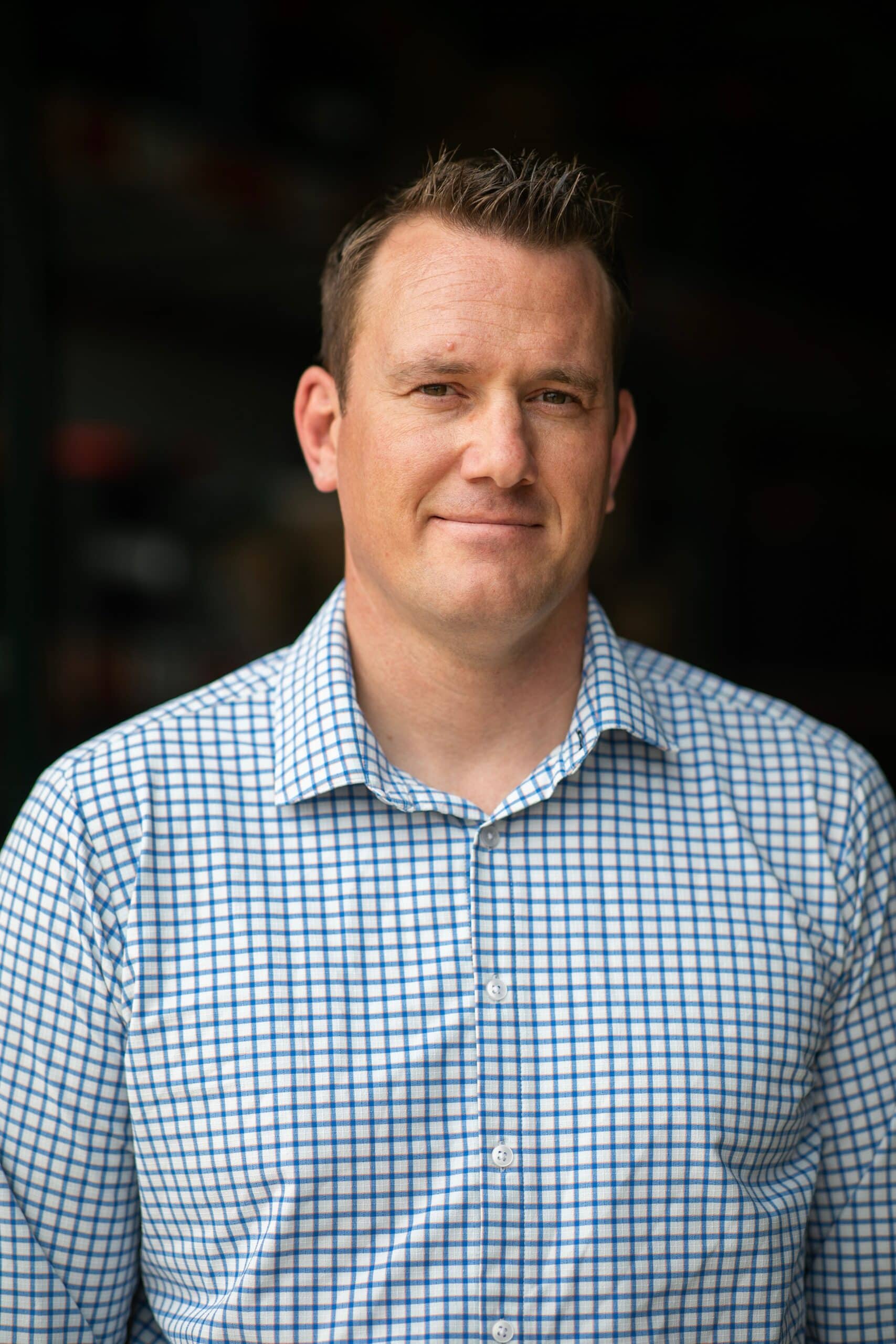 A man with short brown hair wearing a blue and white checkered shirt stands in front of a dark, blurred background, looking at the camera with a neutral expression. - Christopher A. Lighting Design