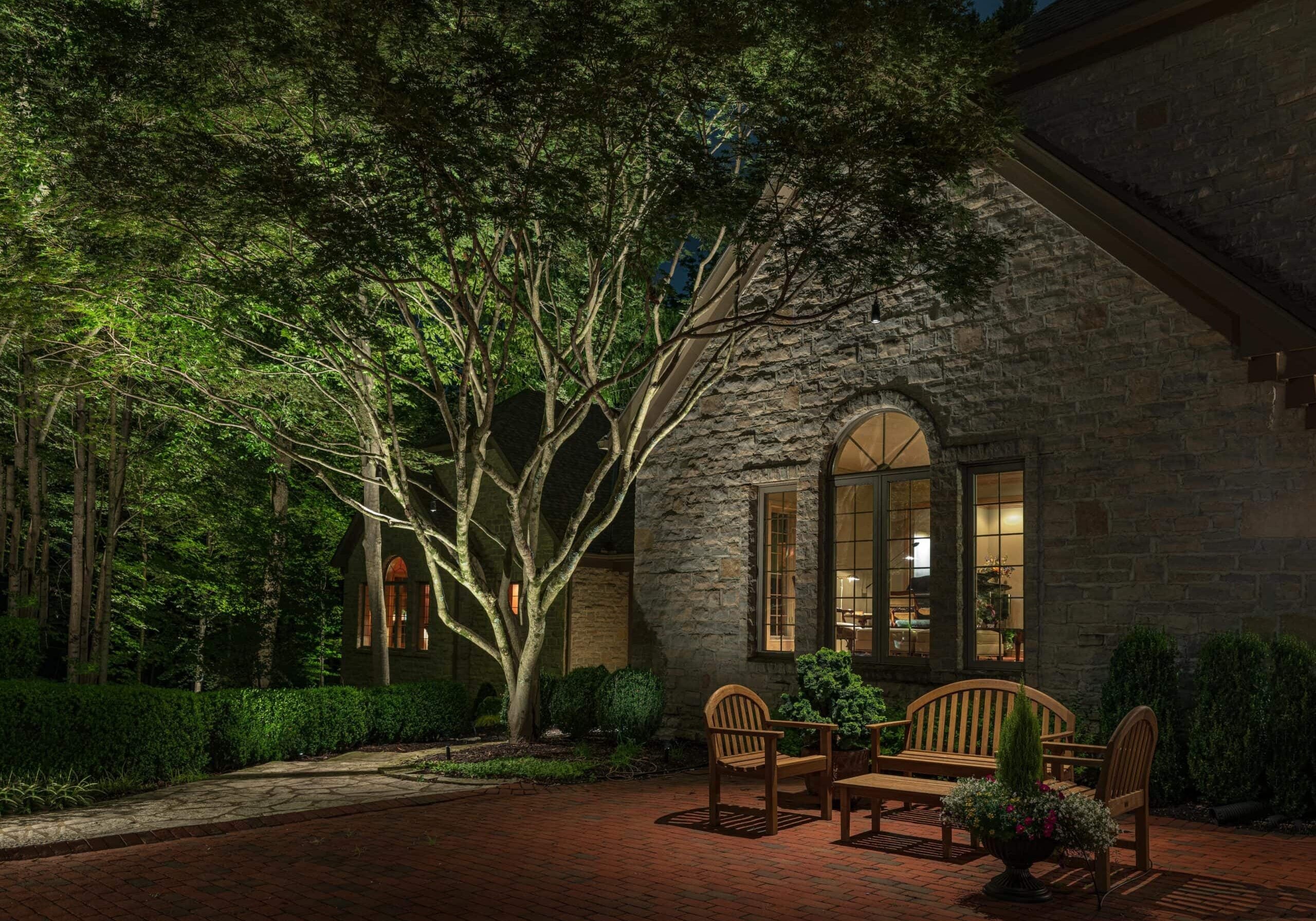 A stone house at night with arched windows, outdoor lights, wooden benches, brick patio, and landscaped greenery and trees.