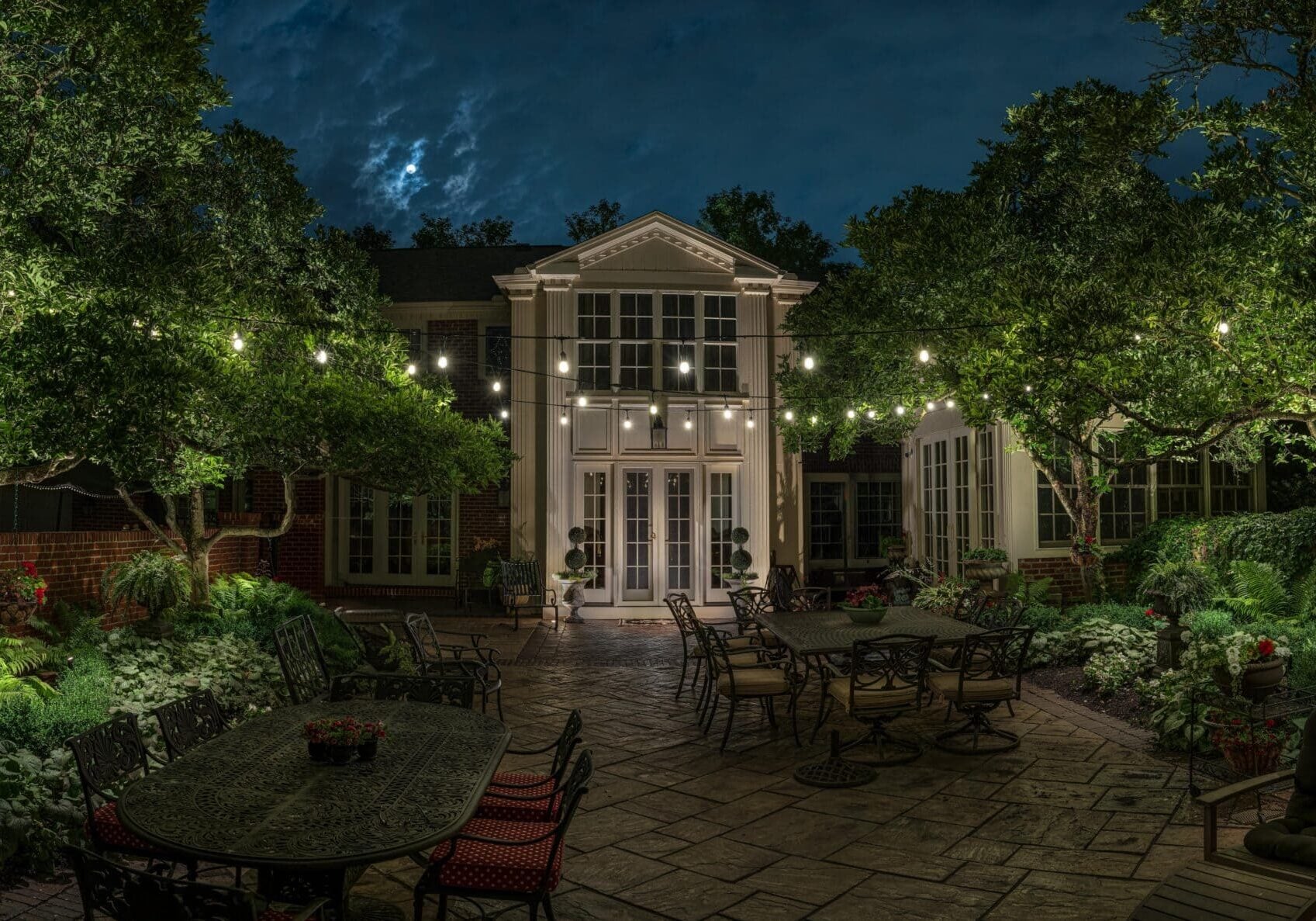Backyard patio at night with string lights, outdoor furniture, lush green plants, and a house with large windows in the background.