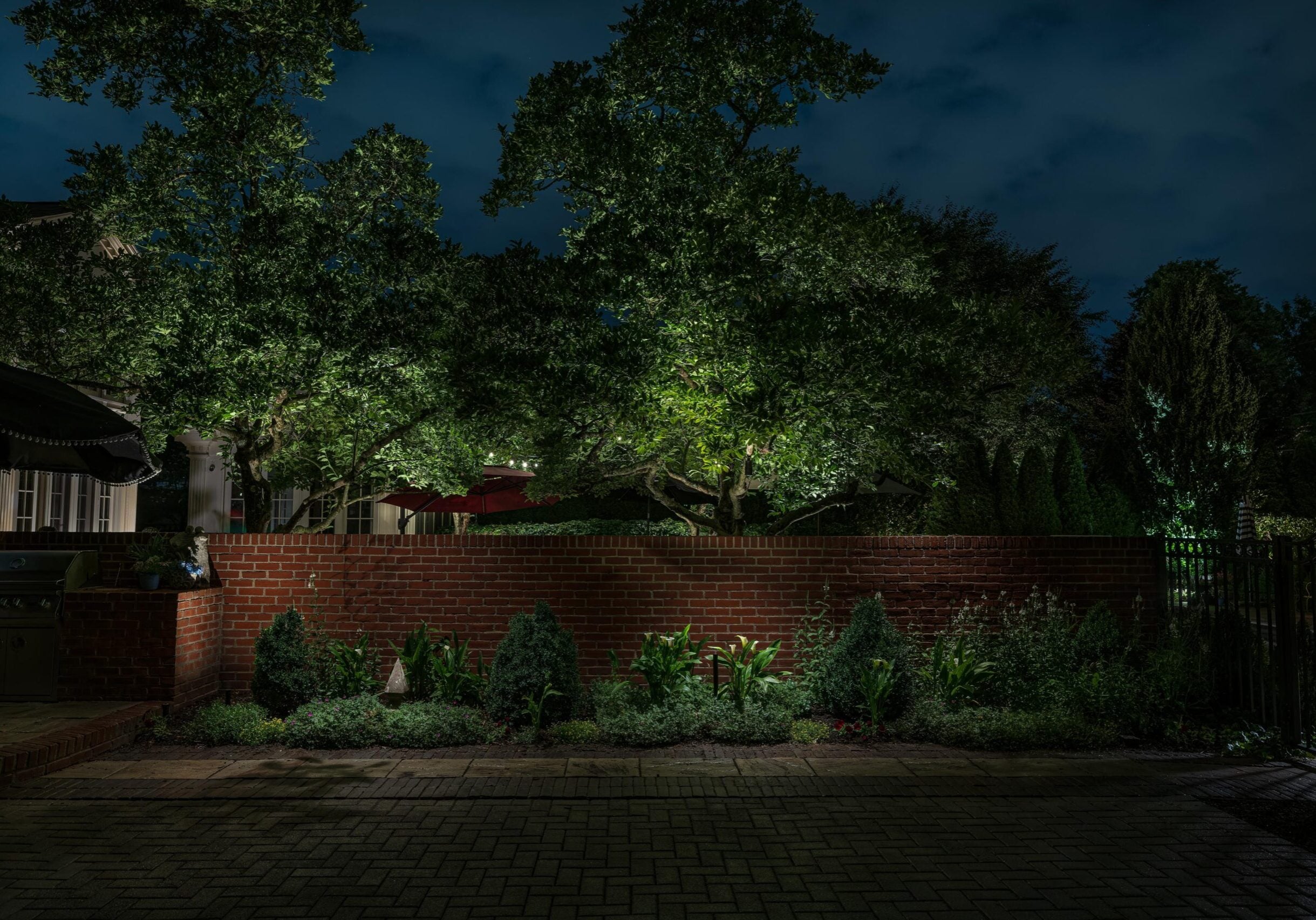A brick patio with a red brick wall and landscaped plants in front, illuminated at night, with large trees and a cloudy sky in the background.