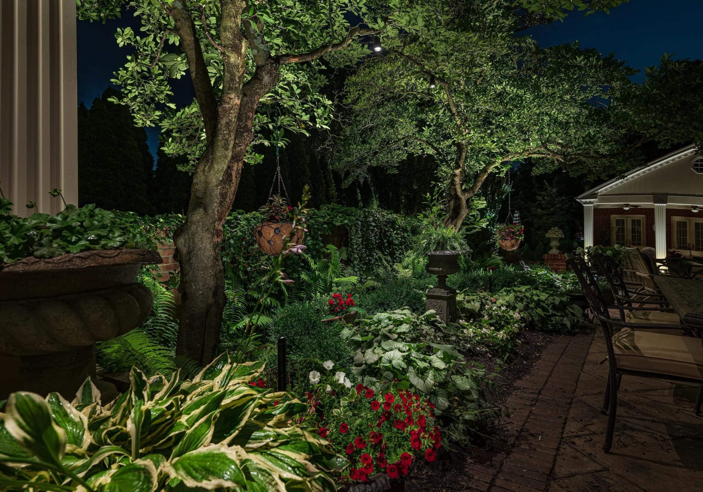 Lush garden at night with trees, flowers, and plants along a brick patio; outdoor furniture and a lit building are visible in the background.