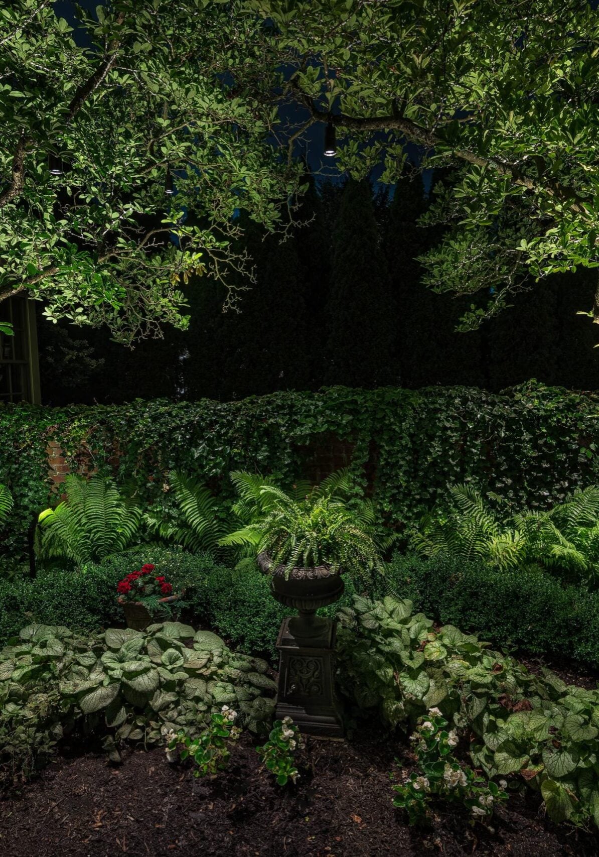 A nighttime garden scene with dense greenery, two large trees, hanging baskets, a variety of plants, and a central birdbath surrounded by foliage.