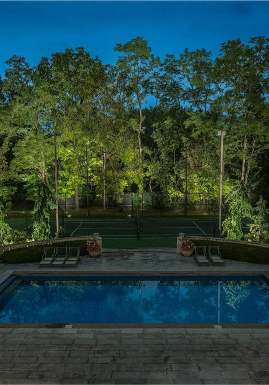 Rectangular swimming pool surrounded by lounge chairs and pavilions, with illuminated tennis court and trees in the background at dusk. - Christopher A. Lighting Design