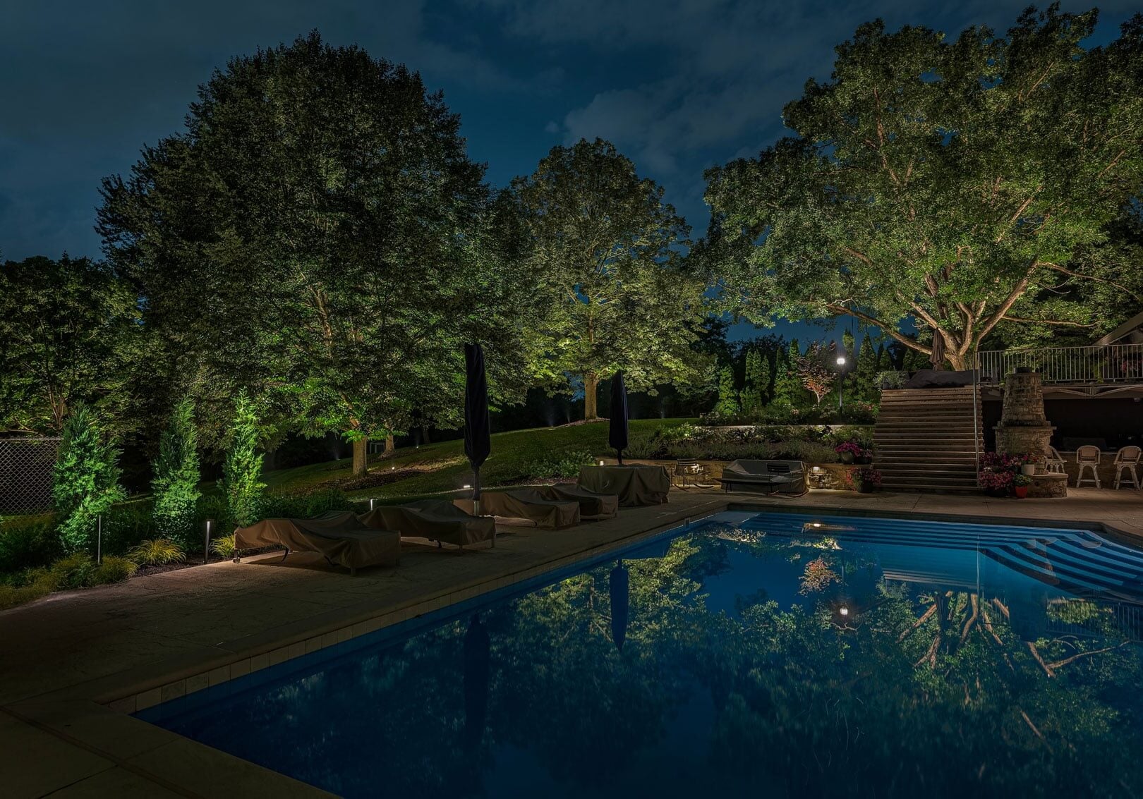 Outdoor swimming pool at night, surrounded by trees and lounge chairs, with lights illuminating the landscape and reflections visible on the water.