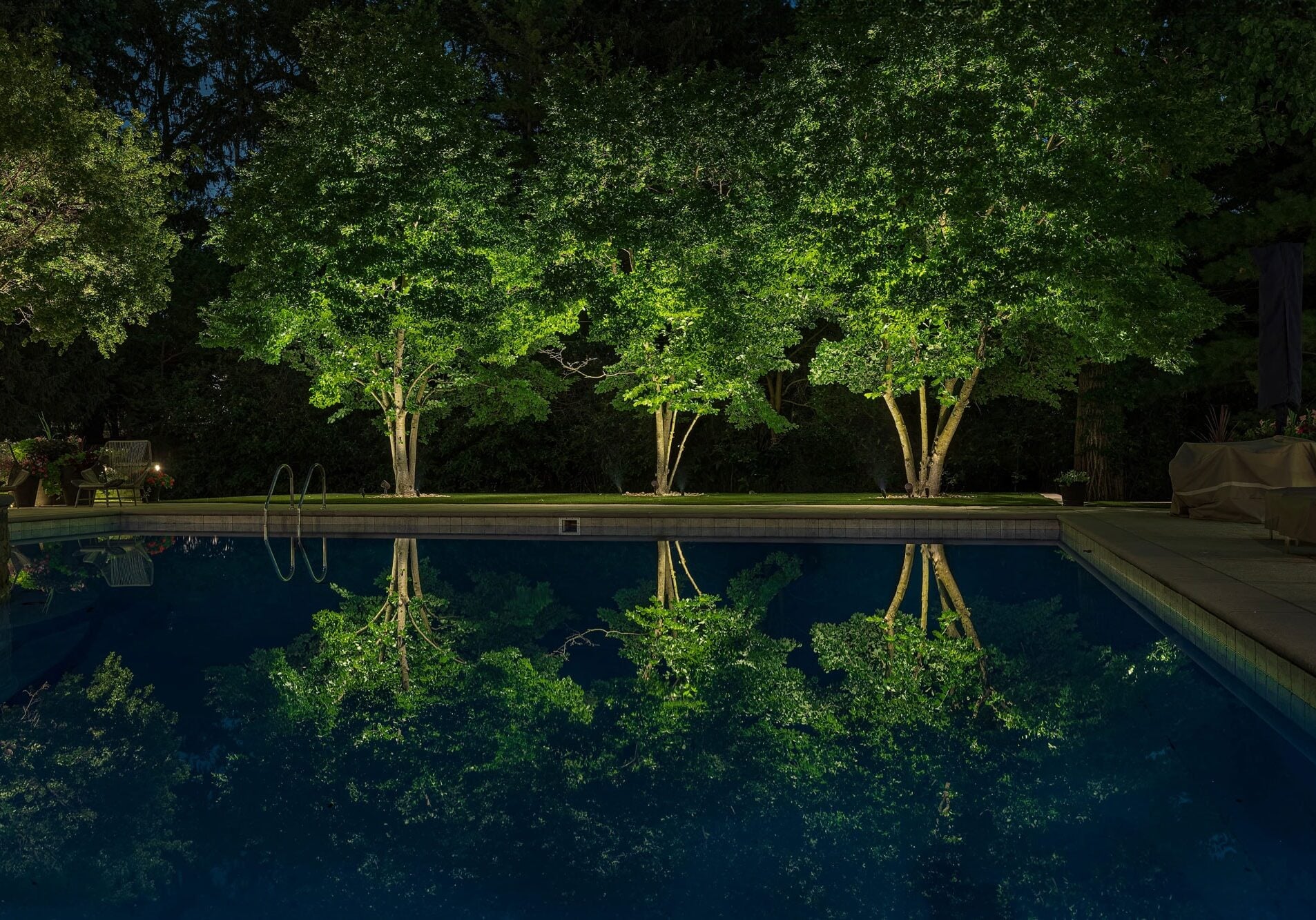 Three illuminated trees are reflected in a swimming pool at night, with surrounding patio furniture visible in the background.