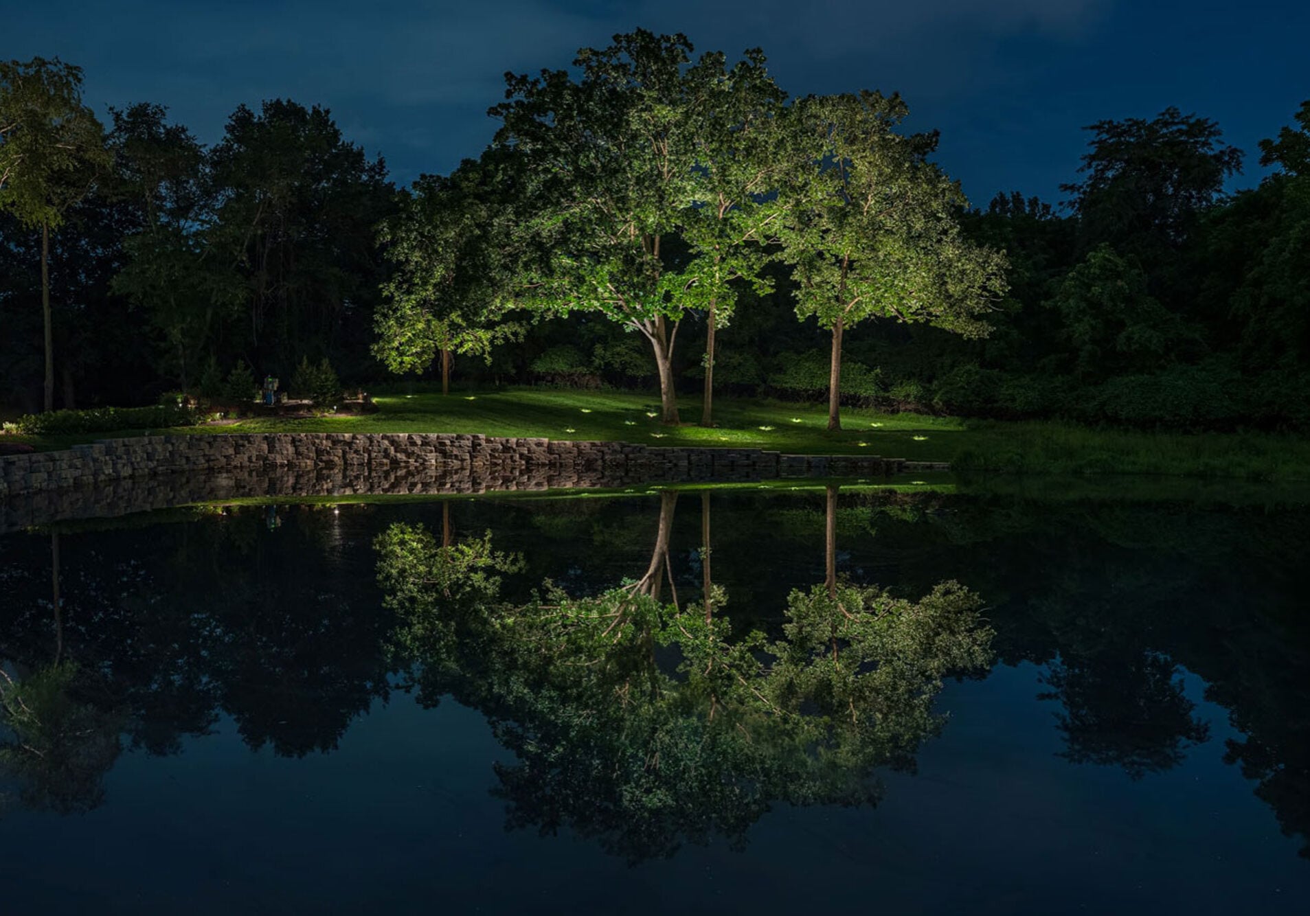 Large trees and grass lit by spotlights are reflected in a calm pond at night, with a stone wall and dense foliage in the background. - Christopher A. Lighting Design