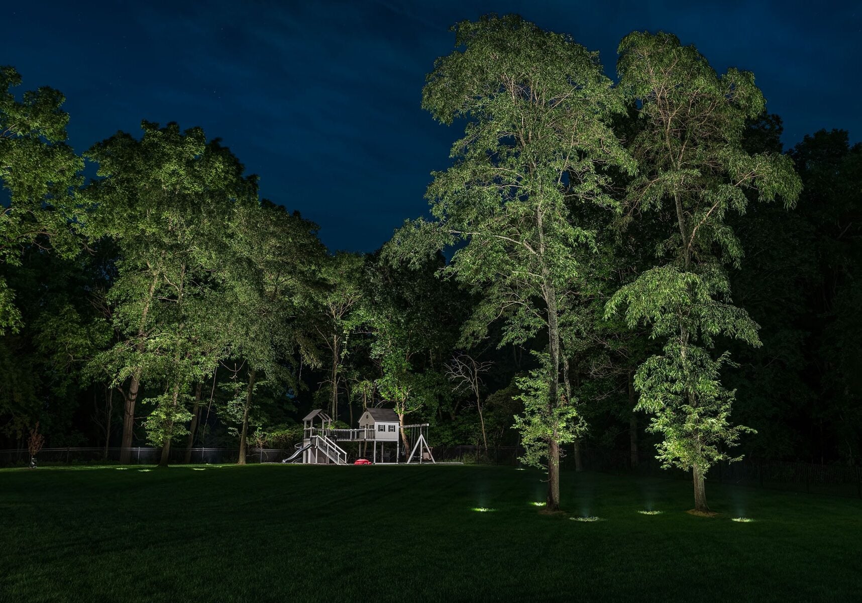 A playground set surrounded by large trees is illuminated at night, with the sky appearing dark blue and the grass in the foreground neatly cut.