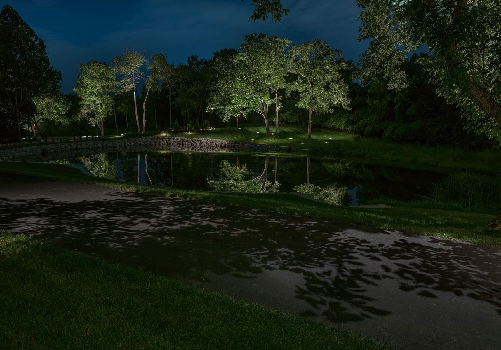 A paved path runs alongside a pond at night, with trees and their reflections visible in the water, all illuminated by artificial lights.