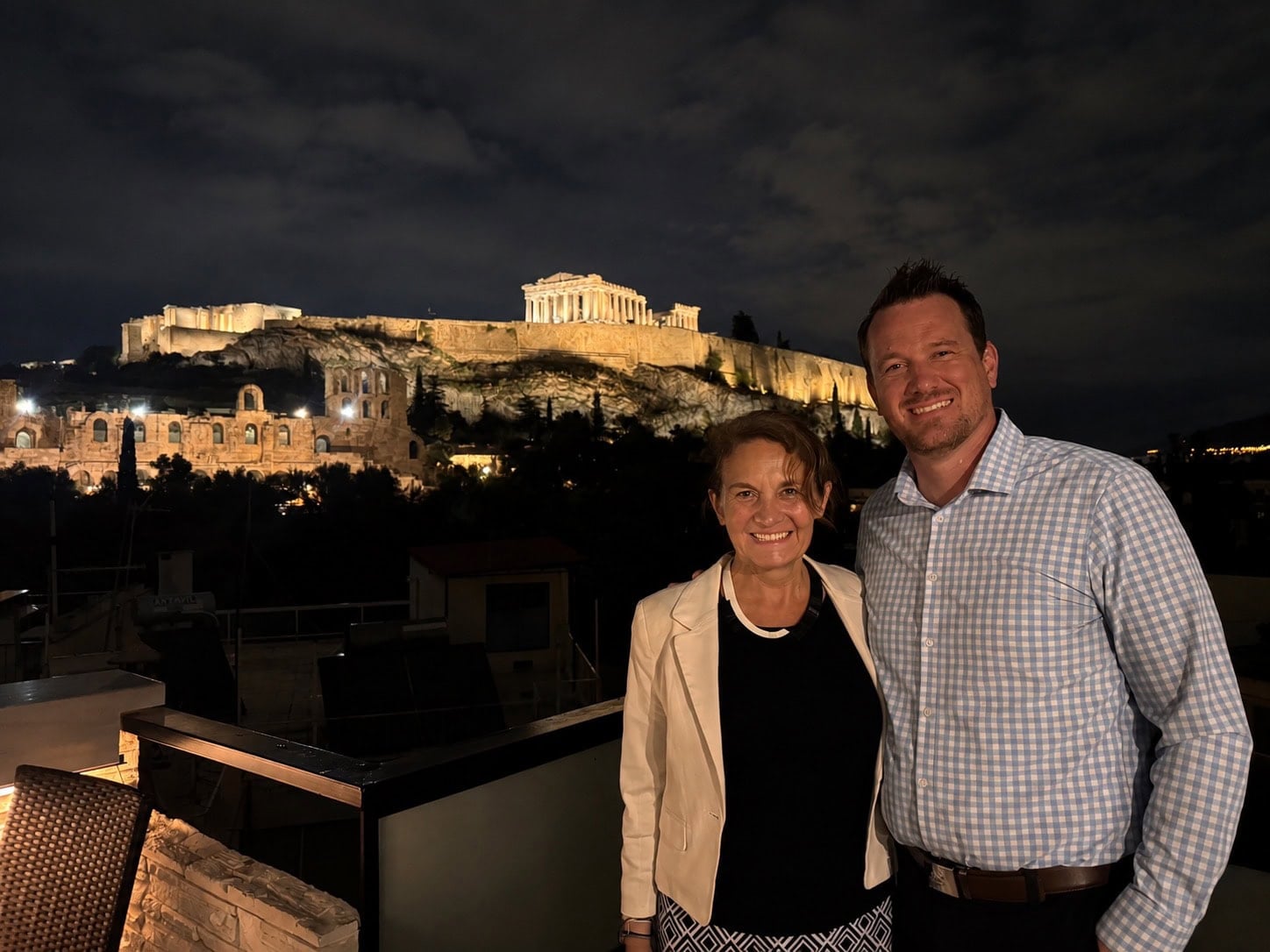 Two people stand and smile on a balcony at night with the illuminated Acropolis and Parthenon in Athens, Greece in the background. - Christopher A. Lighting Design