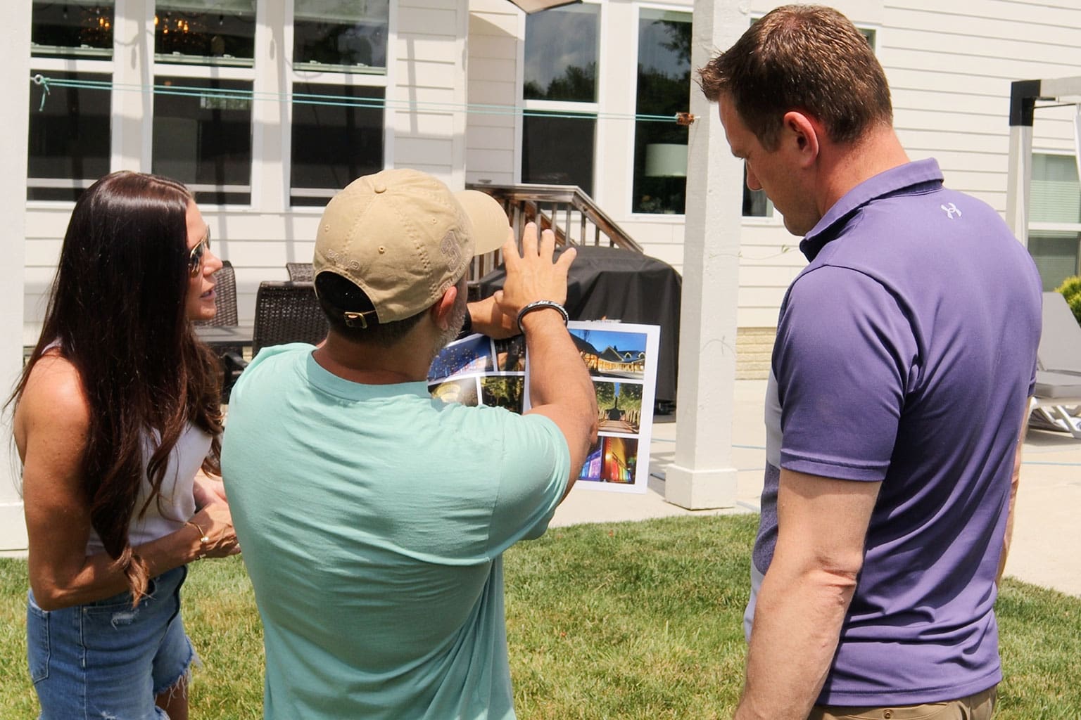 Three people stand on a lawn as one person in a cap shows photos on a sheet, possibly discussing renovation or landscaping plans outside a house. - Christopher A. Lighting Design
