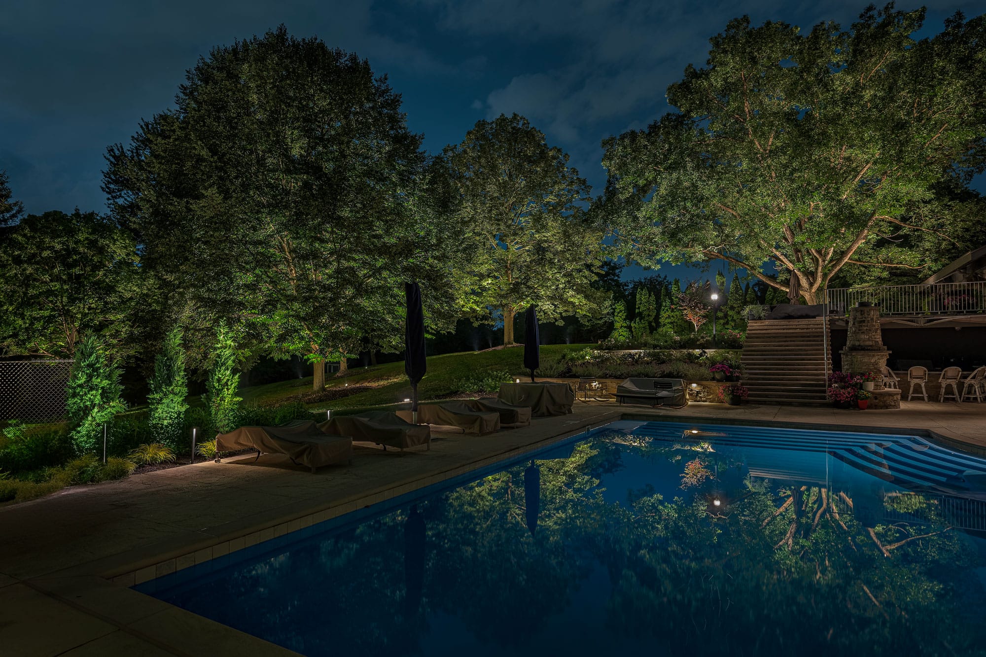A swimming pool at night is surrounded by lounge chairs, large trees, and landscape lights, with reflections visible in the water. - Christopher A. Lighting Design