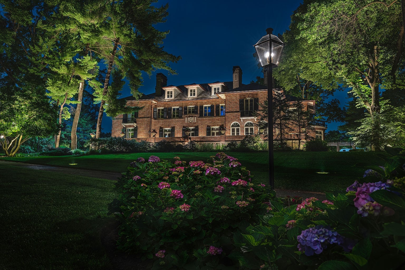 A large brick house stands illuminated at night, surrounded by tall trees, a lamppost, and blooming hydrangea bushes in the foreground. - Christopher A. Lighting Design