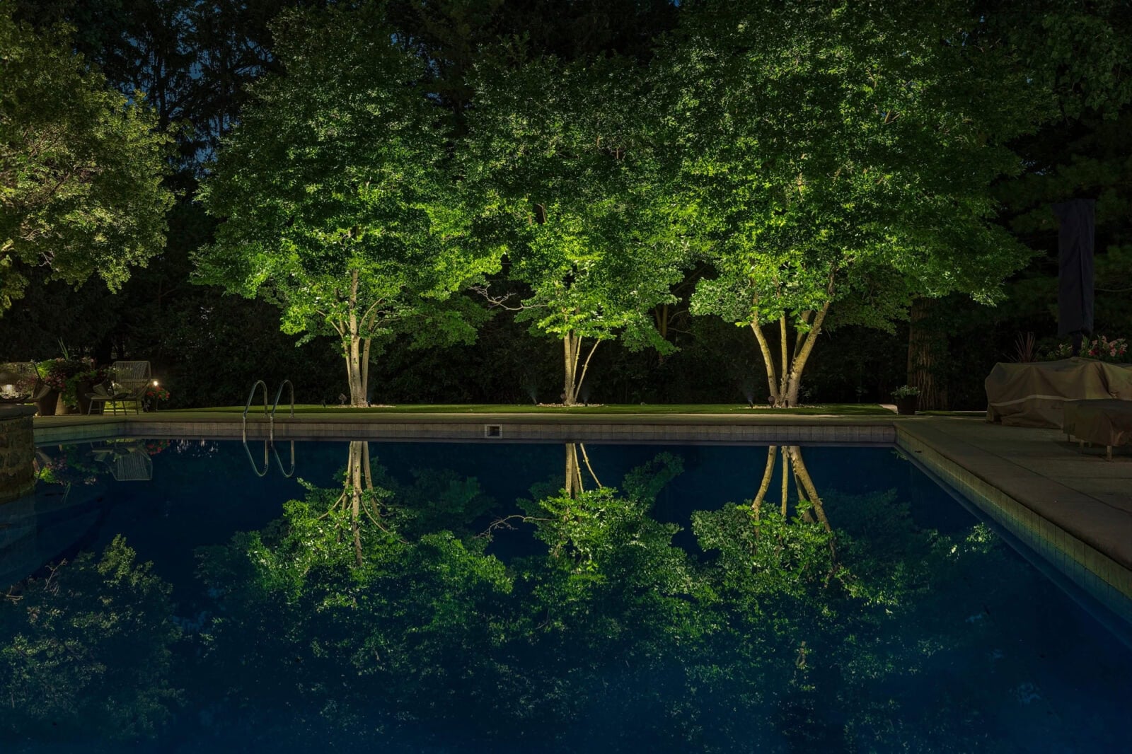 Three illuminated trees are reflected in a swimming pool at night, with surrounding patio furniture visible in the background.