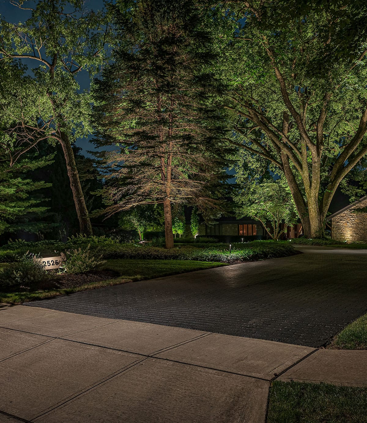 A dimly lit driveway lined with trees and bushes leads to a house at night; a house number sign is visible on the left.