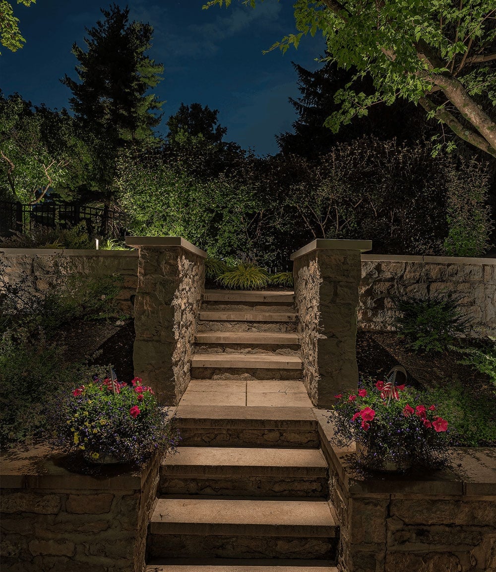 Stone steps with flower pots on either side lead up to a garden terrace, illuminated by outdoor lighting at night, surrounded by trees and plants.