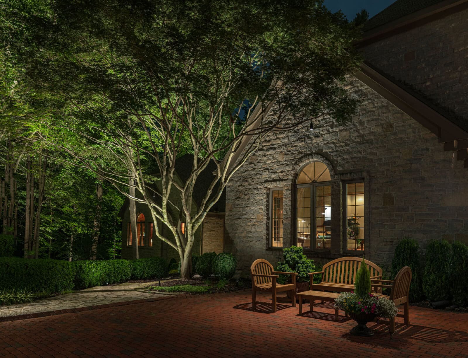 A stone house with arched windows is lit at night, showing wooden benches and potted plants on a red brick patio surrounded by trees and shrubs.