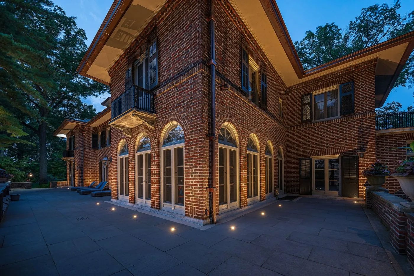 A large two-story brick house with arched windows and exterior lights, surrounded by a spacious paved patio at dusk.