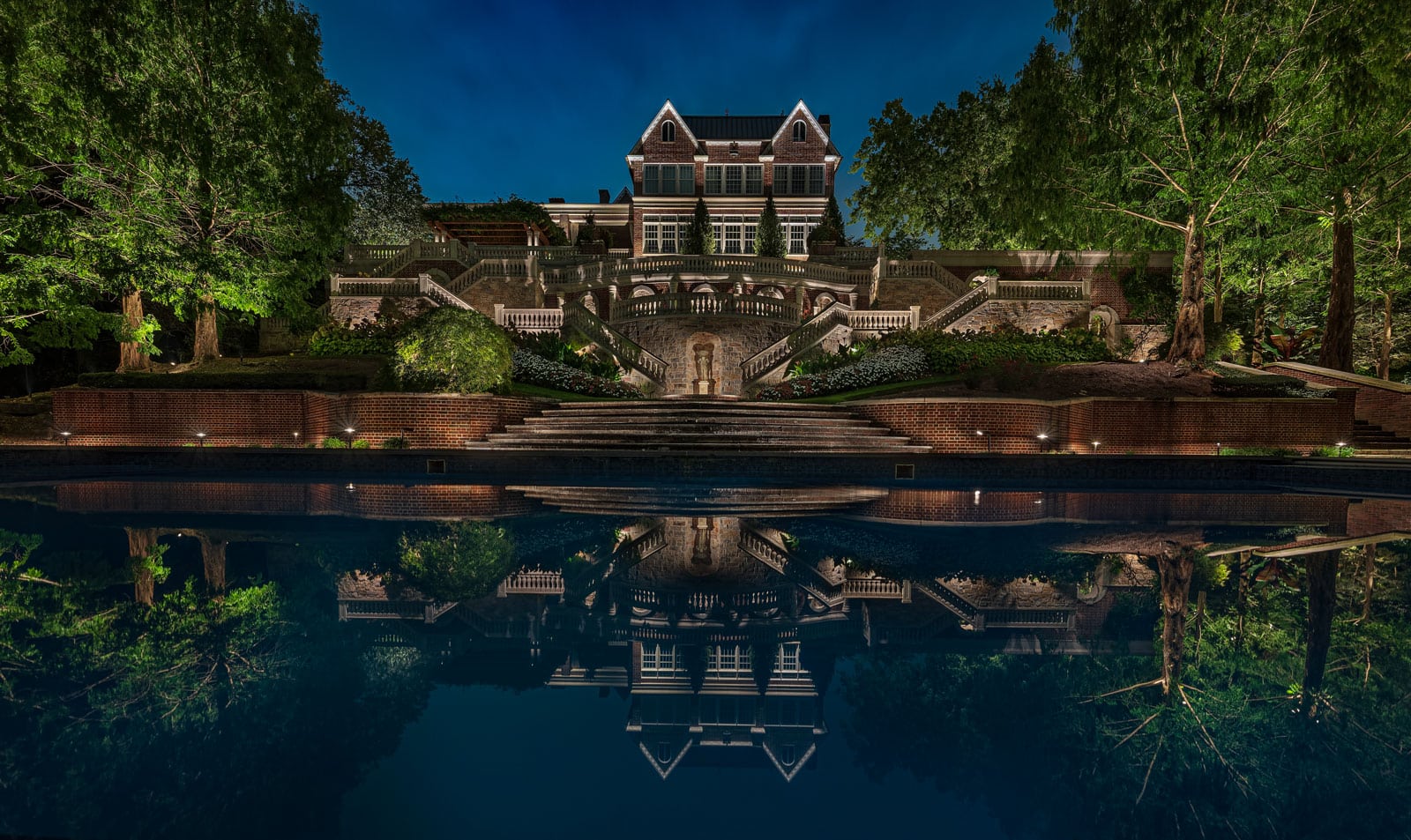 A large brick mansion with multiple balconies and staircases is illuminated at night, reflected in a still pool, surrounded by trees.